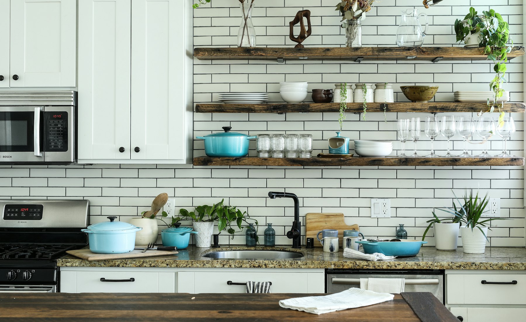 Bright modern kitchen with open wooden shelves holding teal cookware, ceramic bowls and glassware
