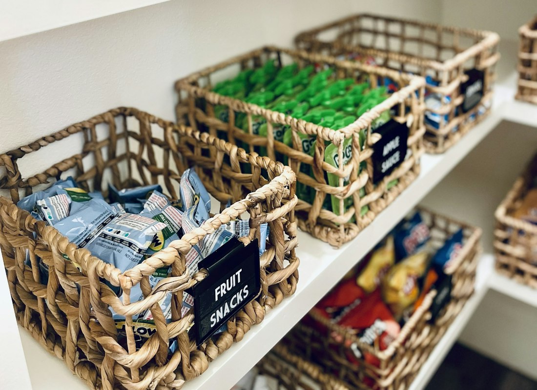 Labelled woven baskets holding snacks on white pantry shelves